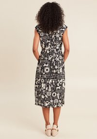 Schwarz gekleidetes Kleid mit weißem Muster, das abstrakte Formen und Tiere zeigt. Ärmelloses Design mit tailliertem Schnitt. Leichte Schuhe ergänzen den Look.