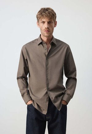 Mann trägt ein braunes Hemd mit Knöpfen, hat die Hände in den Taschen und steht in dunkelblauen Jeans vor einem einfarbigen hellen Hintergrund.