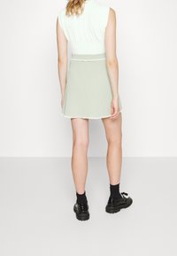 Pletená zelená sukně s rozšířeným tvarem a vlnitým lemem. Má texturovaný povrch. Doplňena černými botami a ponožkami. Neutrální pozadí.