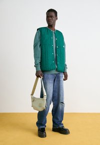 Green quilted vest over a grey sweatshirt, distressed blue jeans, black platform boots, and a beige shoulder bag with a strap.