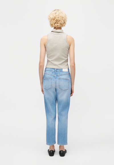 Άτομο με κοντά σγουρά ξανθά μαλλιά που φοράει ένα αμάνικο ριγέ τοπ, ανοιχτόχρωμα μπλε τζιν και μαύρα παπούτσια, που στέκεται με την πλάτη γυρισμένη.