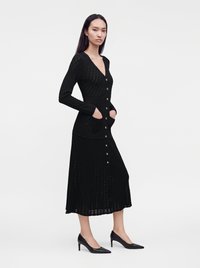 Femme aux longs cheveux foncés portant une longue robe en tricot noire boutonnée avec des poches et des chaussures noires à talons hauts, debout de profil.