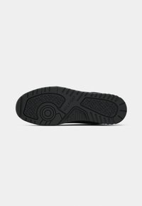 Semelle de chaussure noire vue par le dessous, montrant des motifs de bande de roulement texturés et le nom de la marque "Hilfiger" embossé au centre.