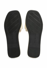 Suela de goma negra con agarre texturizado, material superior beige y acabado suave, que presenta un sutil logo en la zona del talón.