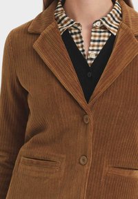 Brauner Cordblazer mit scharfem Kragen, zwei vorderen Taschen und Knopfschluss, über einem schwarzen Weste und einem karierten Hemd getragen.