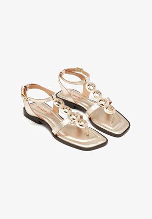 Sandalias metálicas doradas con suela plana, diseño de punta abierta, tira en el tobillo y detalles de acentos metálicos circulares en la parte frontal.