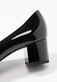 Blank lackpatenterad låg blockklackskor med fyrkantig klack på en vit bakgrund.