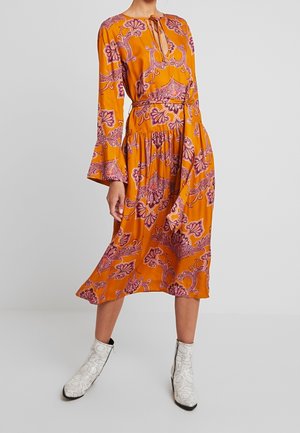 Vrouw draagt een oranje midi-jurk met lange mouwen met roze bloemenpatronen, een keyhole-halslijn, uitlopende manchetten en witte enkellaarsjes.