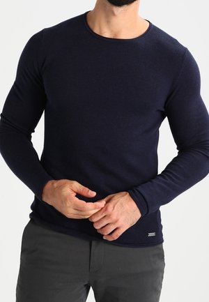 Mand iført en tætsiddende navyblå langærmet jerseytrøje med rund hals og mørke bukser, der justerer sine fingre foran en hvid baggrund.