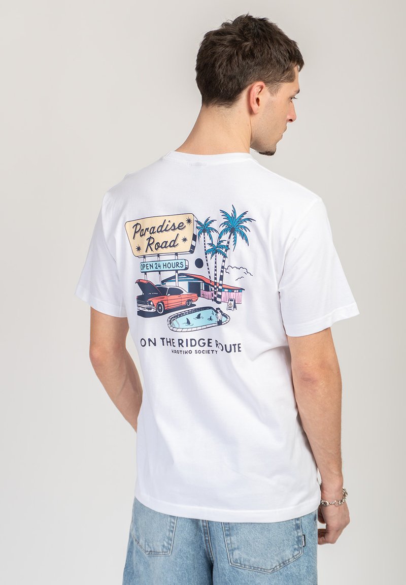 Άνδρας που φοράει λευκό T-shirt με πολύχρωμο vintage αυτοκίνητο, φοινικόδεντρα, πισίνα και γραφικό με την ένδειξη "Paradise Road" στην πλάτη.