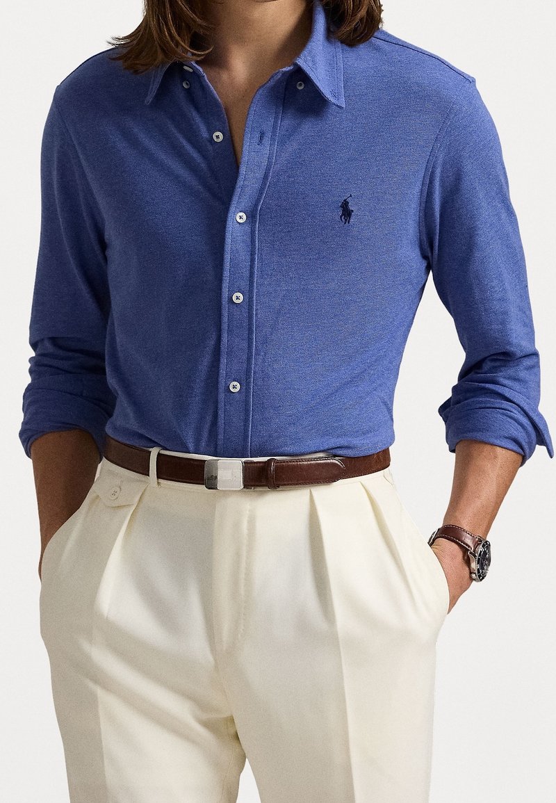 Blå långärmad skjorta med krage i mjukt tyg, med knappar, en logotyp på vänster bröst, i kombination med krämfärgade veckade byxor och ett brunt bälte.