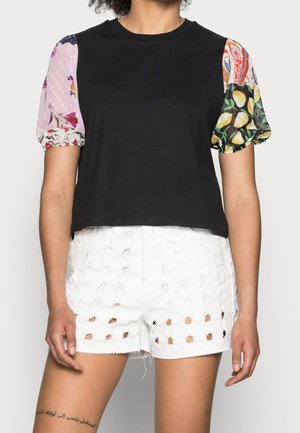 Personne portant un haut noir avec des manches bouffantes fleuries multicolores et un short blanc présentant un motif de découpes carrées.
