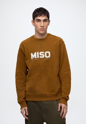 Jeune homme aux cheveux courts et foncés portant un sweat-shirt marron avec le texte "MISO", debout devant un fond blanc uni.
