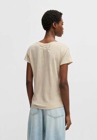 Top beige a maniche corte realizzato in un tessuto leggero, con una leggera arricciatura sul retro e una vestibilità comoda, abbinato a jeans blu chiaro.