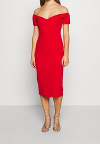Robe midi rouge à épaule dénudée avec une silhouette ajustée et un corsage croisé. Tissu lisse et manches bouffantes courtes, assortie à des talons à lanières.