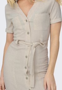 Beige linendress med korte ærmer, nedknapping foran, to brystlommer og en knyttet midjebelte. Strukturert stoff gir visuell interesse.
