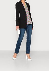 Blazer nero sopra una camicia a fantasia colorata, jeans in denim blu e scarpe col tacco bianche, con una borsa a tracolla elegante in mano.