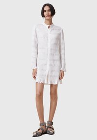 Robe blanche à manches longues avec des rayures textur�ées et un ourlet volanté. Dotée d'un col montant et d'une fermeture à boutons. Portée avec des bottines à motifs.