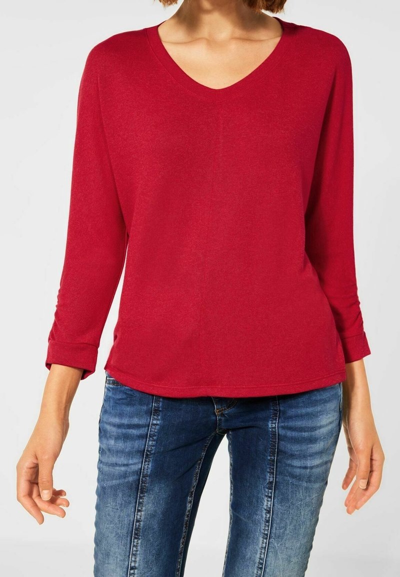 Rotes Langarmshirt mit V-Ausschnitt und entspanntem Schnitt. Hat hochgekrempelte Ärmel und eine glatte Textur. Kombiniert mit blauen Jeans.