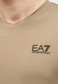 Μπεζ μπλούζα με V λαιμόκοψη που εμφανίζει το λογότυπο EA7 Emporio Armani σε μαύρο στο στήθος, φορεμένη από άτομο με ορατή γραμμή λαιμού και μέρος της γνάθου.