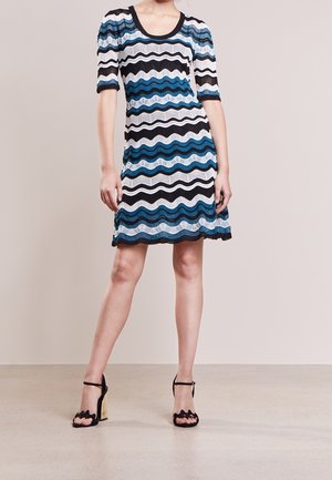 Kvinde iført knælange kjole med sorte, hvide og blå bølgede striber, korte ærmer og sorte høje hæle.