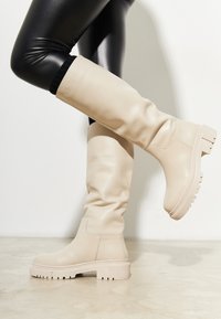 Botine bej până la genunchi, din piele netedă, cu o talpă groasă și texturată, având un design simplu, fără accesorii sau modele vizibile.