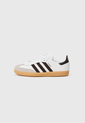 Hvid Adidas Samba sneaker med sorte striber, gummisål, beige tåhætte og sort hælflap, vist fra siden på en lys baggrund.