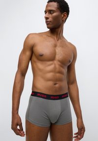 Gray boxer briefs with a black waistband featuring red "HUGO" logo. The fabric appears smooth and fitted, accentuating the muscular physique.