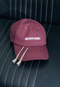 Casquette de baseball ajustable bordeaux avec "NEVER QUIET." brodé en blanc, avec deux pinces à cheveux argentées à côté sur une surface noire texturée.