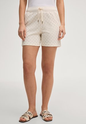 Frau trägt hellbeige gemusterte Shorts mit Kordelzugbund und beige J O O P! Slide-Sandalen vor neutralem Hintergrund.