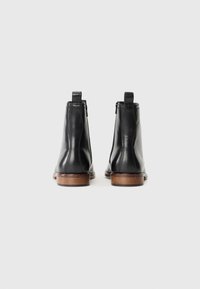 Un par de botas de cuero negro hasta el tobillo con suelas de madera marrón, vistas desde la parte trasera sobre un fondo blanco liso.
