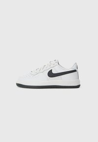 Bijele niske Nike tenisice s crnim swoosh logom, crnom potplatom i bijelim vezicama na jednostavnoj svijetlo sivoj pozadini.