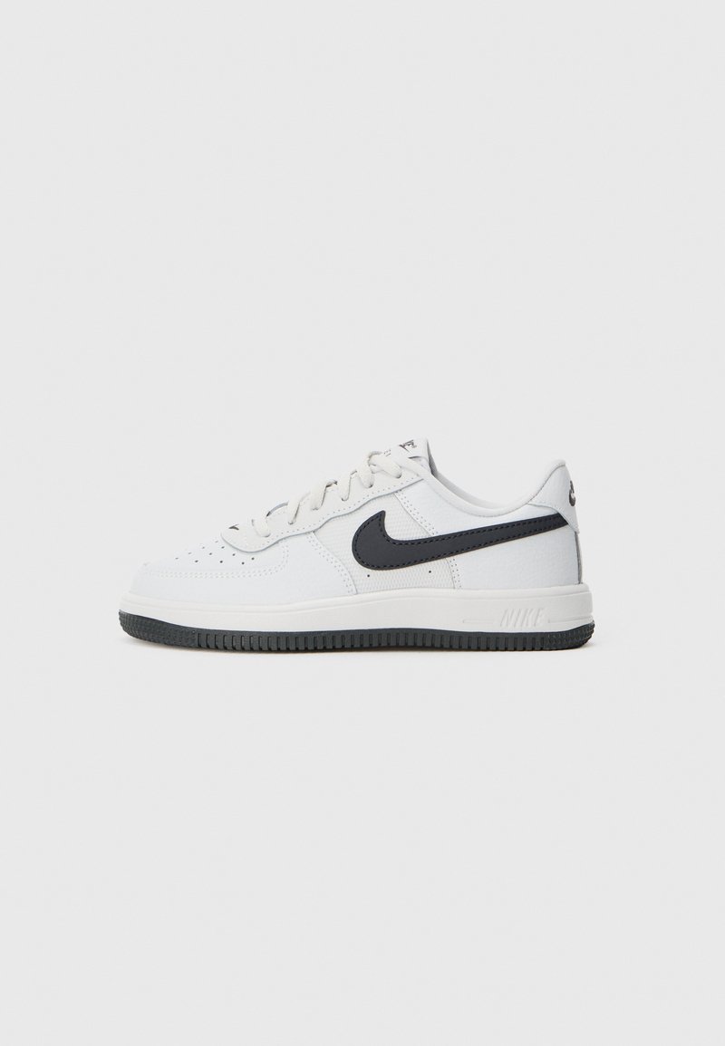 Bijele niske Nike tenisice s crnim swoosh logom, crnom potplatom i bijelim vezicama na jednostavnoj svijetlo sivoj pozadini.