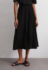 A person stands wearing a black pleated midi skirt and black slide sandals, with a textured light gray background.