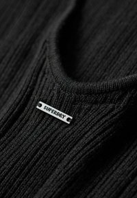 Svart ribbad tyg med rund halsringning. Har en rektangulär svart tagg med "SUPERDRY" präglat i vitt. Texturerade ytdetaljer synliga.