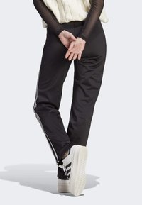 Pantalones de chándal negros con rayas laterales blancas, hechos de una tela suave, con un diseño ajustado. Combinados con zapatillas blancas y suela texturizada.