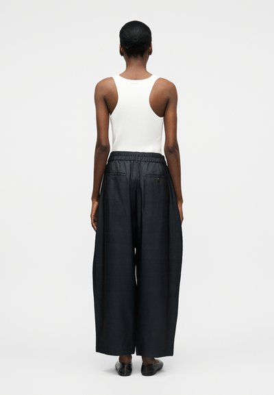 Άτομο με σκουρόχρωμο δέρμα και κοντά μαλλιά, φορώντας λευκό αμάνικο τοπ και φαρδιά μαύρη παντελόνια, γυρισμένο πλάτη σε απλό ανοιχτόχρωμο φόντο.