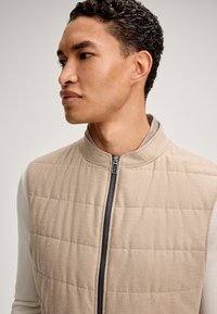 Beige gesteppte Weste mit hohem Kragen, Reißverschluss und weicher Textur. Die Ärmel haben eine kontrastierende helle Farbe. Einfaches und modernes Design.