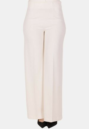 Pantaloni larghi color crema a vita alta indossati con scarpe nere a punta, mostrati dalla vita ai piedi su uno sfondo semplice.