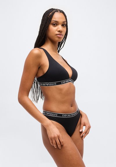 Γυναίκα με μακριά πλεξούδες που φοράει μαύρο σουτιέν και εσώρουχα Emporio Armani, στέκεται μπροστά σε ένα απλό φωτεινό φόντο.