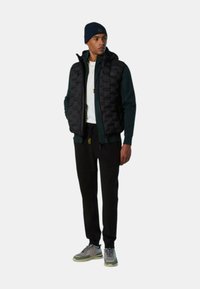 Chaleco acolchado negro sobre sudadera verde oscura, camisa blanca, pantalones jogger negros y zapatillas grises con toques verdes. Un gorro completa el conjunto.