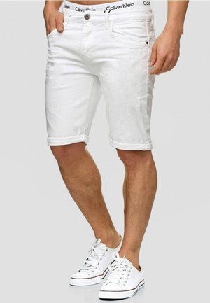 Mężczyzna w białych spodenkach z podwiniętym brzegiem, widoczna gumka w talii z logo Calvin Klein, oraz białe niskie trampki z sznurowadłami związanymi na kokardę.