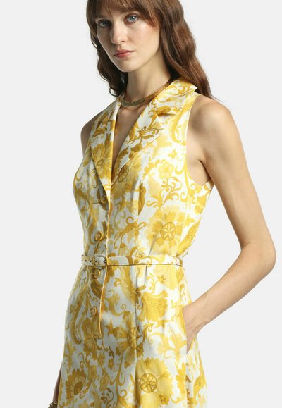 Ung kvinde iført hvid ærmeløs kjole med gult blomsterprint og matchende bælte, stående med den ene hånd i lommen.