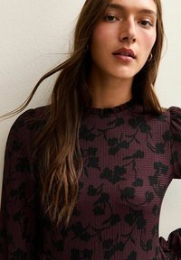 Maglione a maglia testurizzata bordeaux con motivo floreale nero, caratterizzato da un colletto arricciato e maniche a sbuffo. Finitura liscia con accenti eleganti.