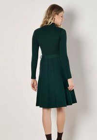 Robe en maille vert foncé, avec une texture côtelée, un col montant et une jupe évasée avec des plis. Détail de taille noué à l'arrière.