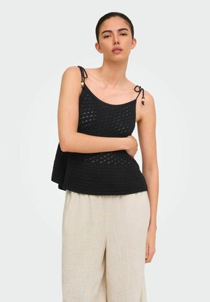 Femme portant un haut sans manches en maille noire avec des bretelles nouées aux épaules et un pantalon beige ample, debout devant un fond clair uni.