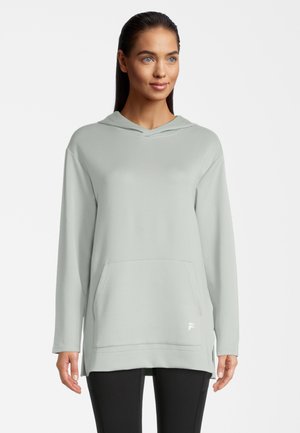 Vrouw in een lichtgrijze hoodie met een buidelzak en zwarte leggings, lichtjes naar rechts gekeerd op een effen witte achtergrond.