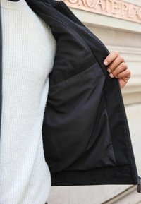 Schwarze Jacke mit einem Reißverschluss auf der Vorderseite, die ein glattes, schwarzes Innenfutter und eine Tasche zeigt. Das Außenmaterial wirkt strukturiert.