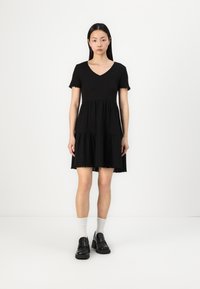 Black, short-sleeved dress with a V-neck, tiered design, and frayed hem. Worn with black shoes and white ankle socks.