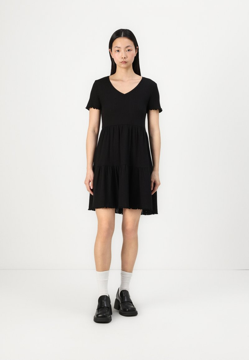 Black, short-sleeved dress with a V-neck, tiered design, and frayed hem. Worn with black shoes and white ankle socks.
